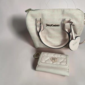 May include: A light pink Juicy Couture handbag and matching wallet set. The handbag features a top handle, a shoulder strap, and the brand name in gold lettering. The wallet is quilted with a heart-shaped clasp.
