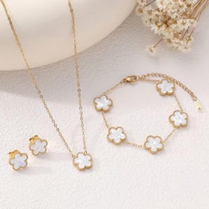 Könnte beinhalten: Ein goldfarbenes Schmuckset mit Halskette, Armband und Ohrringen. Jedes Schmuckstück hat ein kleeblattförmiges Design mit weißem Mittelteil. Das Set wird auf einer weißen Oberfläche mit getrockneten Blumen im Hintergrund präsentiert.