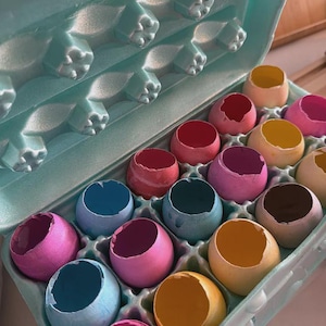 May include: An open, light blue egg carton filled with colorful, empty eggshells. The shells are painted in various shades, including pink, blue, yellow, and brown. The carton is made of a recycled material.