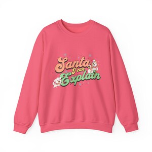 Puede incluir: Una sudadera rosa brillante con mangas largas. La parte delantera presenta el texto "Santa I can Explain" en una fuente retro, junto con ilustraciones de un muñeco de nieve y un perro. El diseño incluye copos de nieve y estrellas, creando un tema festivo navideño.