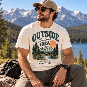 Puede incluir: Una camiseta de manga corta color crema con un gráfico de montañas, pinos, un lago y un sol naranja. El texto dice OUTSIDE Is a Good IDEA y EXPLORE MORE WORRY LESS. La prenda se combina con pantalones verde oliva y una gorra de béisbol color canela.