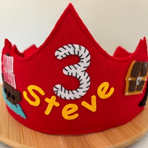 May include: A red felt birthday crown with the number "3" in white and the name "Steve" in yellow. The crown has decorative elements including a treasure chest and a ship. The crown is placed on a wooden plate.