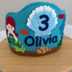 May include: A turquoise felt crown with a mermaid, seashell, and fish design. The crown features the number "3" and the name "Olivia" in navy blue. The base is brown.