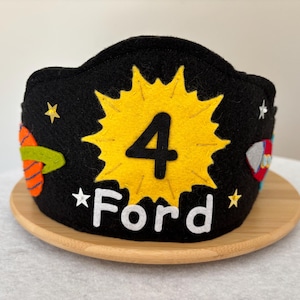 May include: A black felt birthday crown with a yellow number 4 in a starburst design. The name "Ford" is below the number. The crown is decorated with stars and colorful space-themed appliqués, sitting on a wooden plate.