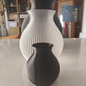 Op de afbeelding: Een verzameling van drie decoratieve vazen in verschillende maten en kleuren. De grootste vaas is lichtgrijs, de middelste is zwart en de kleinste is zwart. Alle vazen hebben een geribbelde textuur en een afgeronde vorm.