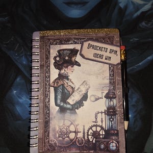 May include: A spiral-bound notebook featuring a graphic of a person in a top hat holding a document, with gears and a lamp below. The text reads SPROCKETS SPIN, IDEAS WIN. A black pencil with a pink eraser is attached to the side of the notebook.