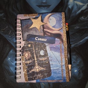 May include: A spiral-bound notebook featuring a collage design with a gold star, a crescent moon held by a hand, and a label reading Cosmic. The cover includes celestial imagery, a sun and moon scale illustration, and gold glitter accents with a black pencil.
