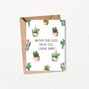 May include: A birthday greeting card with a kraft paper envelope. The card features a white background with watercolor illustrations of cacti in pots. The text reads: "Another year older, you're still lookin' sharp!"