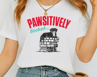 Può includere: T-shirt bianca con la scritta "PAWSITIVELY Booked..." in rosso e verde acqua. La maglietta presenta un'illustrazione in bianco e nero di un cane seduto su una pila di libri. Un capo casual per tutti i giorni.