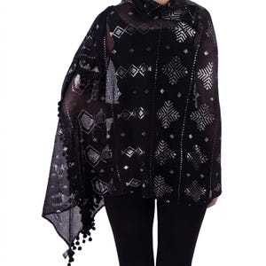 May include: Black shawl with silver geometric patterns and small pom-poms along the edge. The sheer fabric drapes over the shoulders, creating a stylish accessory. The design includes diamond and snowflake-like shapes.