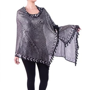 May include: A black and silver poncho with a geometric pattern. The poncho has a cowl neck and bell sleeves. The edges are trimmed with black pom-poms. The fabric has a shimmering effect.