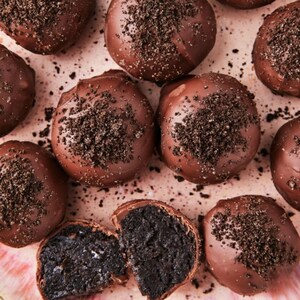 May include: An overhead shot showcases several chocolate-covered truffles, some whole and others cut open to reveal a dark interior. The truffles are sprinkled with dark crumbs, resting on a light pink surface.