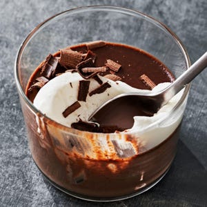 May include: A glass of chocolate pudding topped with whipped cream and chocolate shavings. A silver spoon is partially submerged in the dessert, ready to take a bite. The pudding is a rich, dark brown color.
