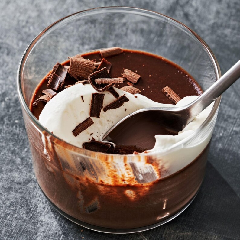 May include: A glass of chocolate pudding topped with whipped cream and chocolate shavings. A silver spoon is partially submerged in the dessert, ready to take a bite. The pudding is a rich, dark brown color.