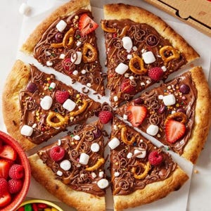 May include: A dessert pizza cut into slices, topped with chocolate, pretzels, marshmallows, sprinkles, and fresh berries. The pizza crust is golden brown, and the toppings are arranged on a white surface.