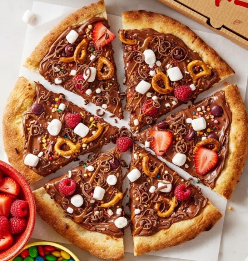 May include: A dessert pizza cut into slices, topped with chocolate, pretzels, marshmallows, sprinkles, and fresh berries. The pizza crust is golden brown, and the toppings are arranged on a white surface.