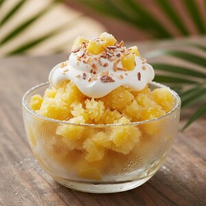 May include: A clear glass bowl filled with yellow shaved ice, topped with whipped cream, diced pineapple, and toasted coconut flakes. The dessert sits on a wooden surface, with blurred green palm leaves in the background.