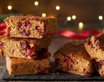 May include: A stack of square-cut, golden-brown baked goods with visible red fruit pieces, possibly cranberry. The treats are arranged on a dark slate board, with a wooden background and blurred string lights, suggesting a festive theme.