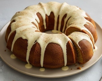 May include: A golden-brown bundt cake with a creamy white glaze drizzled over the top. The cake is on a beige plate, and a slice has been cut out. The glaze is lightly dusted with a spice.