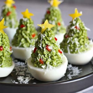 Puede incluir: Huevos rellenos decorados como árboles de Navidad. El relleno verde está adornado con trozos de pimiento rojo, queso rallado y una estrella amarilla. Un aperitivo festivo para las fiestas.