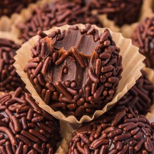 May include: Close-up of several chocolate truffles, each coated in chocolate sprinkles and nestled in a paper cup. One truffle has a bite taken out of it, revealing the soft, chocolatey interior. The truffles are a rich brown color.