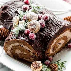 May include: A chocolate Yule log cake, decorated with sugared cranberries, powdered sugar, and small meringue mushrooms. The cake is on a white platter, with pine cones and sprigs of rosemary.