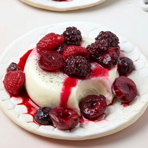 May include: A white panna cotta dessert topped with fresh red raspberries, blackberries, and cherries, drizzled with a vibrant red sauce. The dessert is served on a white plate with a honeycomb pattern.