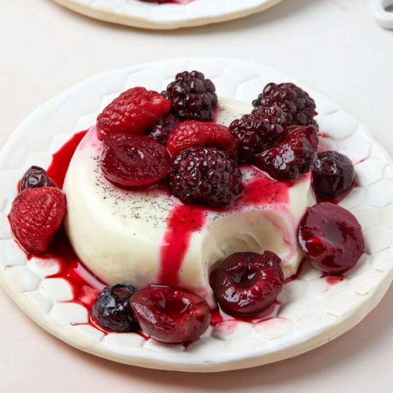 May include: A white panna cotta dessert topped with fresh red raspberries, blackberries, and cherries, drizzled with a vibrant red sauce. The dessert is served on a white plate with a honeycomb pattern.