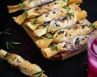 May include: A wooden cutting board displays several baked pastries wrapped in golden-brown paper, garnished with sprigs of rosemary and black sesame seeds. The pastries are arranged on a dark surface, with a glass of red liquid visible in the corner.