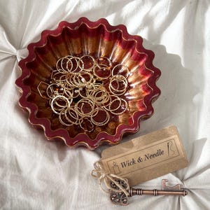 Puede incluir: Un cuenco decorativo con borde festoneado en burdeos y dorado contiene numerosos anillos dorados. Una etiqueta con el texto "Wick & Needle" y una llave decorativa están junto al cuenco. El cuenco está sobre una superficie blanca texturizada.
