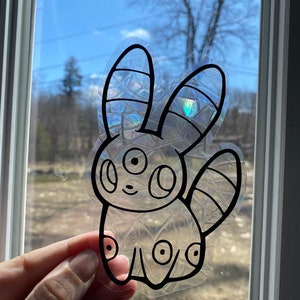May include: A clear, iridescent sticker of a cartoon character with large ears, three eyes, and a tail. The character is outlined in black.