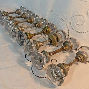 May include: A collection of six clear glass door knobs with an octagonal shape, each featuring a metallic base and a central threaded metal spindle. The knobs are arranged in a row, showcasing the transparent faceted glass and aged metal hardware.