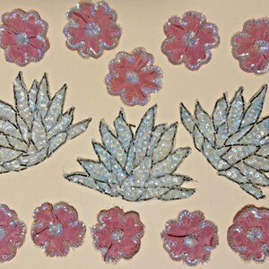 May include: A collection of fabric appliques featuring five pink flowers with sequin centers and edges, and three white leaf-shaped clusters embellished with iridescent sequins and silver thread outlines, arranged on a light beige surface.