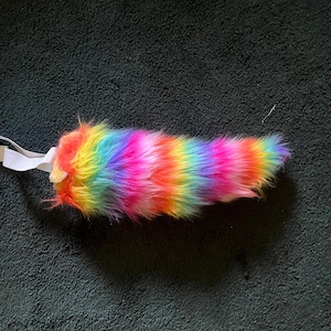 May include: A plush, faux fur tail featuring a vibrant, multi-colored rainbow pattern with horizontal stripes of red, orange, yellow, green, blue, and purple. The accessory includes a white elastic band attached to one end for secure fastening.