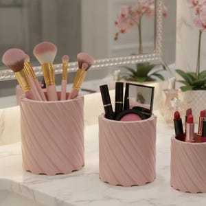 Op de afbeelding: Een verzameling roze make-up kwasten houders en cosmetica containers op een marmeren aanrecht. De houders hebben een textuur met een wervelend ontwerp en bevatten make-up kwasten, lippenstiften en andere schoonheidsproducten. Een spiegel en decoratieve bloemen zijn op de achtergrond.