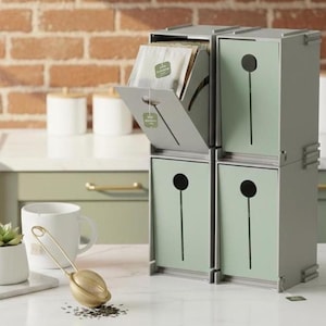 Pode incluir: Um organizador de saquinhos de chá de quatro compartimentos num esquema de cores verde claro e cinzento. Cada compartimento tem uma pega circular e uma ranhura vertical. Uma caneca branca, um infusor de chá dourado e uma pequena suculenta num vaso branco também são visíveis.