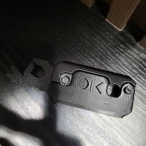 Puede incluir: Una herramienta negra impresa en 3D con las letras "OK" en la superficie. La herramienta tiene un cuerpo rectangular con bordes redondeados y una extensión en forma de llave en un lado. La herramienta está sobre una superficie oscura.