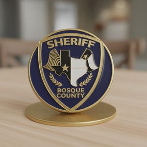 May include: A circular gold-toned metal badge featuring a dark blue enamel inlay. The centre displays a map of Texas with a star, flanked by flag motifs. Text reads SHERIFF at the top and BOSQUE COUNTY at the bottom, mounted on a gold base.