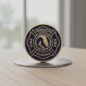 May include: A circular navy blue and gold metal challenge coin featuring the Charlotte County Fire EMS emblem. The design includes a Maltese cross, the text Charlotte County Florida, the year 1921, and a central map of Florida with a fish icon.