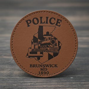 May include: A circular, brown leather coaster with the word "POLICE" at the top. The centre shows a black graphic of the state of Maryland with a train and the text "BRUNSWICK MD 1890".