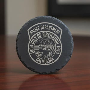 May include: A round, dark gray slate coaster with the words "POLICE DEPARTMENT CITY OF FIREBAUGH CALIFORNIA" in white lettering. The coaster features a detailed emblem with a river, sun, and the years 1854 and 1914.