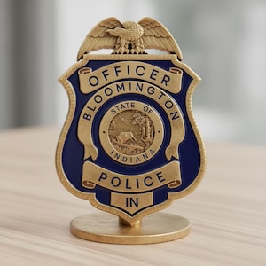 May include: A gold-colored police badge with a blue shield shape. The badge features the words "OFFICER BLOOMINGTON POLICE IN" and an eagle at the top. The center of the badge has a circular emblem with a deer.