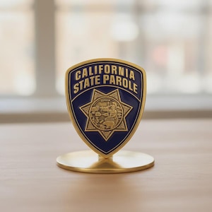 May include: A gold-colored metal desk ornament featuring a blue shield with the words "CALIFORNIA STATE PAROLE" and a gold star emblem. The ornament is mounted on a circular base.