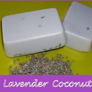 May include: Two bars of white soap with lavender buds embedded in the soap. The soap is sitting on a yellow surface with dried lavender buds scattered around the soap.