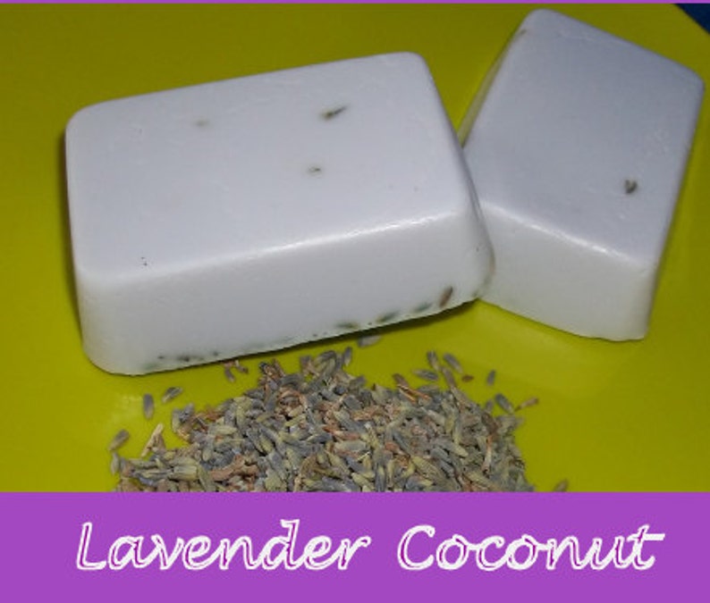 May include: Two bars of white soap with lavender buds embedded in the soap. The soap is sitting on a yellow surface with dried lavender buds scattered around the soap.