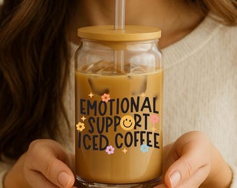 May include: A clear glass jar with a wooden lid and straw, filled with iced coffee. The jar has the text "EMOTIONAL SUPPORT ICED COFFEE" with flower and smiley face graphics. The jar is held by a person.