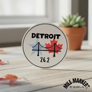 May include: A round sticker with the word "DETROIT" in black, above a bridge graphic and the number "26.2". The bridge is flanked by a blue star and a red maple leaf. The sticker is white with a black border.