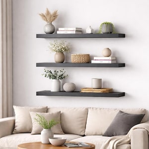 Può includere: Tre mensole fluttuanti grigio scuro montate su una parete bianca, ciascuna con oggetti decorativi. Le mensole espongono vasi con fiori secchi, libri, un cestino intrecciato e piante in vaso. Sotto c'è un divano beige con cuscini.