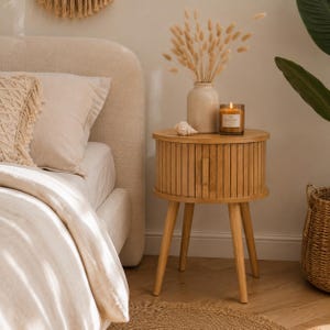 Può includere: Un comodino in legno marrone chiaro con un piano rotondo e un singolo cassetto. Il comodino ha tre gambe angolate. Una candela accesa in un barattolo di vetro, una conchiglia e un vaso con fiori secchi sono sopra. Il comodino è accanto a un letto.
