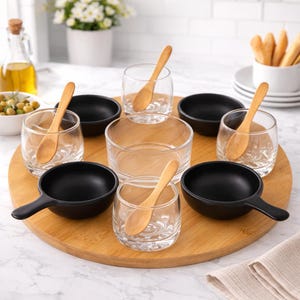 Puede incluir: Una bandeja de madera presenta pequeños cuencos negros, vasos transparentes y cucharas de madera. Un cuenco de vidrio transparente está en el centro. El conjunto está sobre una superficie de mármol blanco.